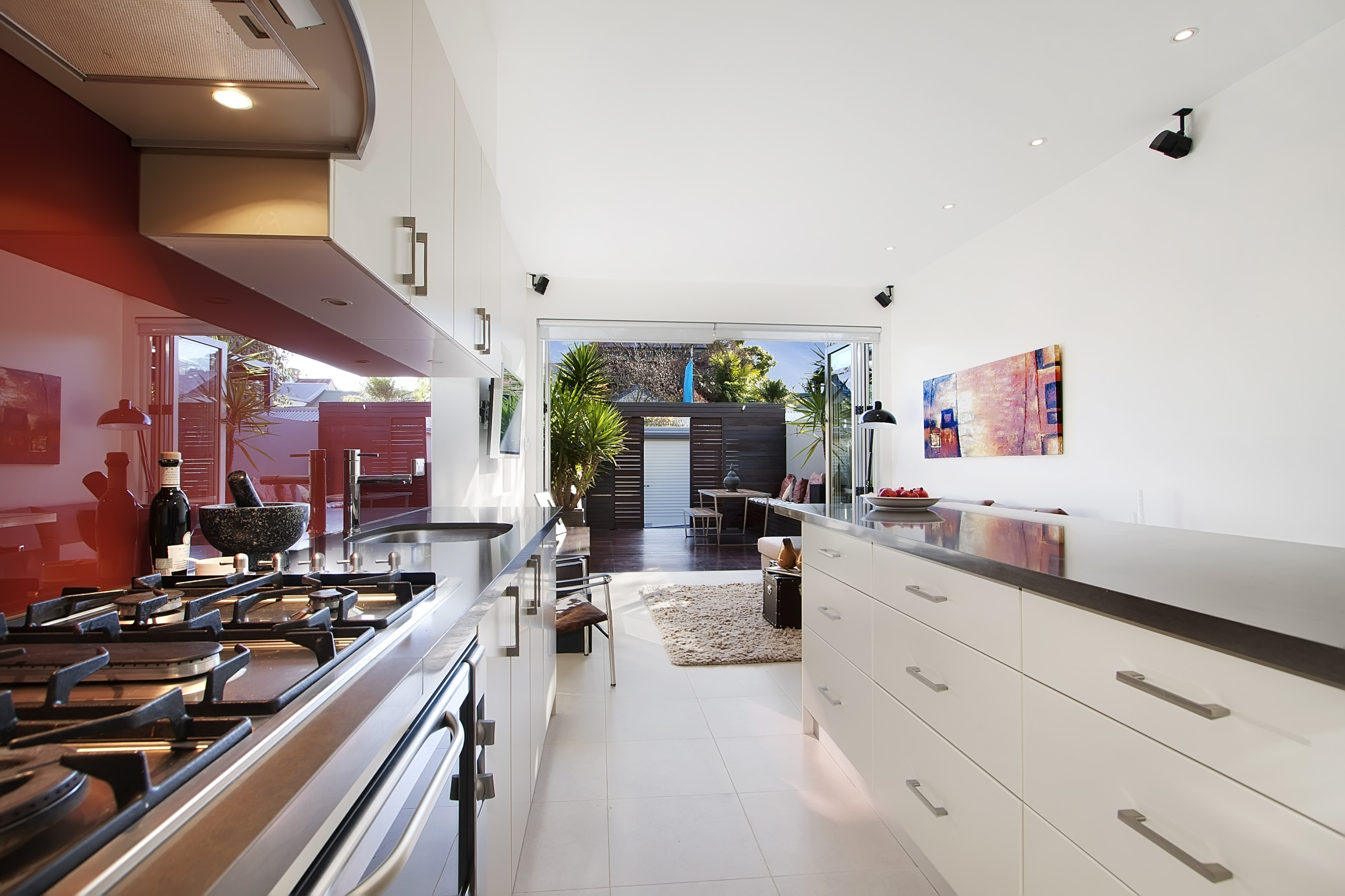 White kitchen with red splash back, featuring outdoor timber fencing