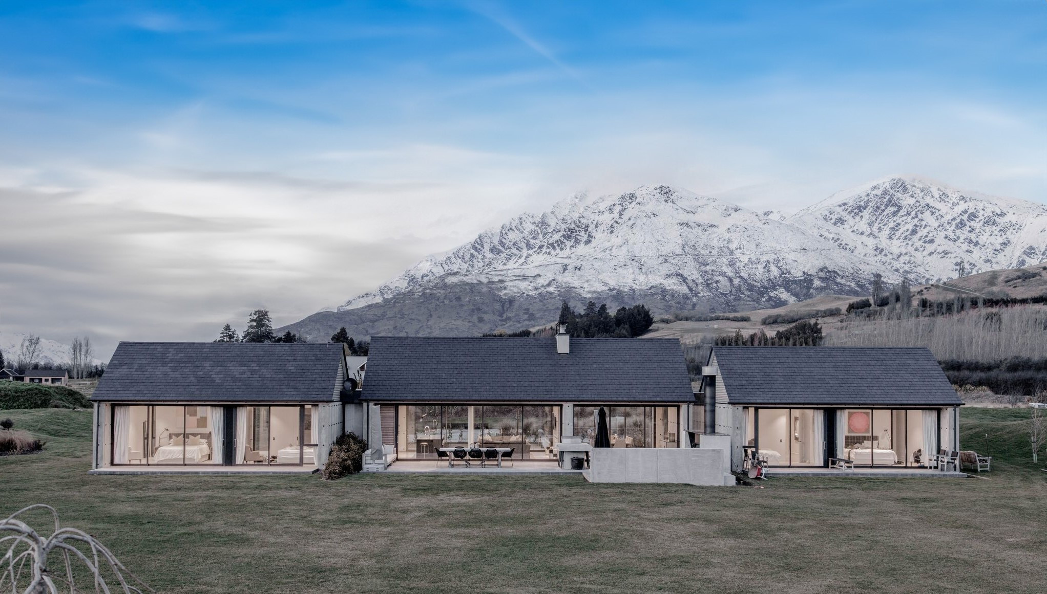 A modern house with mountains in the background, featuring wall-to-ceiling windows and doors