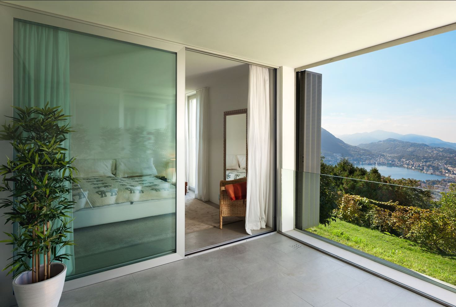 Glass sliding doors opening to a bedroom with a deck and glass balustrade, offering panoramic views of the ocean and mountains